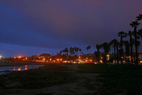 Evening view of a beach with palm trees and street lamps lit up along a boardwalk under a twilight sky.