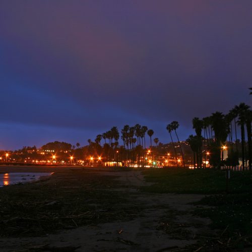 Evening view of a beach with palm trees and street lamps lit up along a boardwalk under a twilight sky.