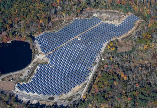 Aerial view of a large solar farm next to a lake, surrounded by a forest with autumn foliage colors.