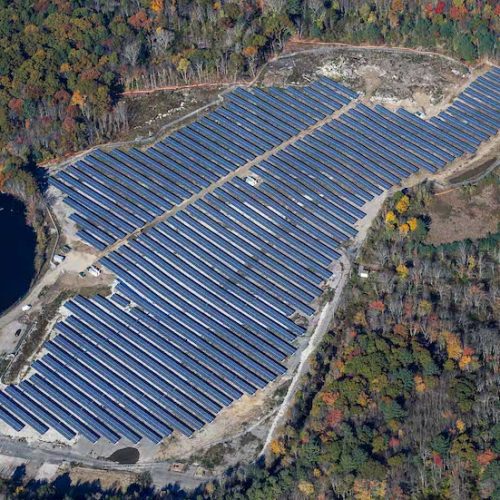 Aerial view of a large solar farm next to a lake, surrounded by a forest with autumn foliage colors.