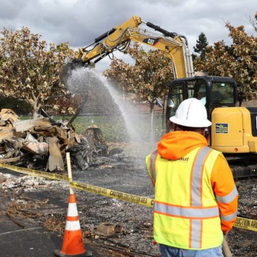A construction worker in a high-vis vest watches an excavator spray water on demolition debris surrounded by caution tape and cones.