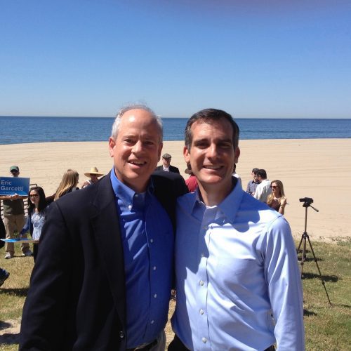 Two men smiling for a photo on a sunny beach with others and a camera in the background. one wears a blue suit and the other a blue shirt.