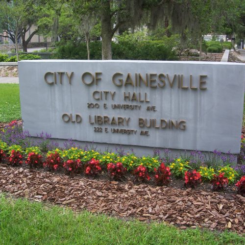 A sign in front of gainesville city hall with addresses for city hall and the old library building, surrounded by colorful flowers.