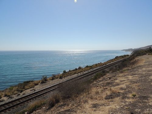 Railroad tracks running parallel to a coastline with a clear blue ocean under a sunny sky.