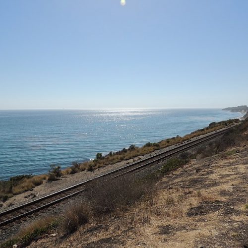 Railroad tracks running parallel to a coastline with a clear blue ocean under a sunny sky.
