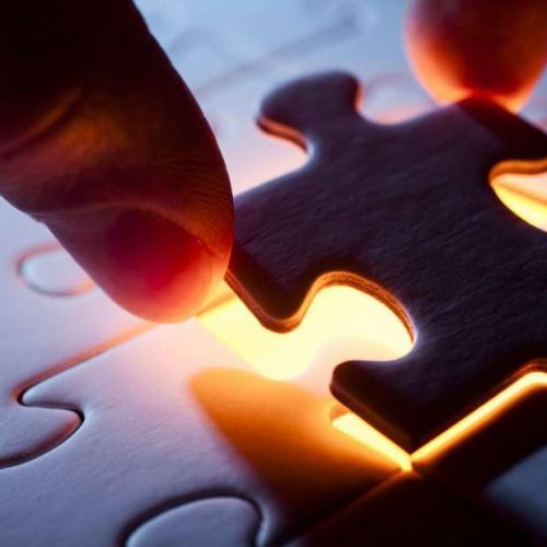 A person's fingers placing the last piece into a jigsaw puzzle, which glows from beneath on a wooden surface.