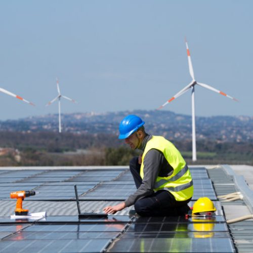 Engineer in safety gear inspecting solar panels with wind turbines in the background on a sunny day.