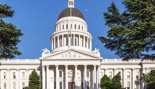 Exterior view of the california state capitol building in sacramento, framed by lush green trees under a clear blue sky.