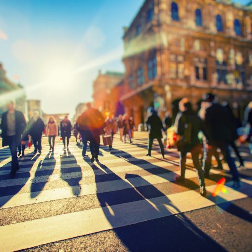 Blurred image of pedestrians crossing a city street, with sunlight creating lens flares in the background.