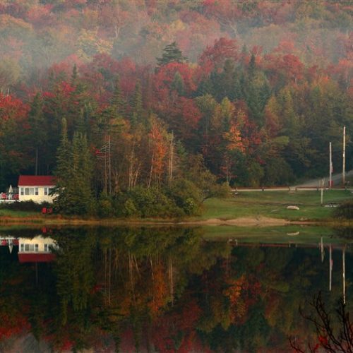 A tranquil autumn landscape featuring a placid lake reflecting colorful trees, with a small white house and a boat nestled among the foliage.