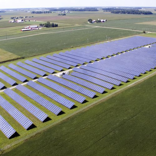 Aerial view of a large solar farm with rows of solar panels amidst green fields, with farm buildings in the background.