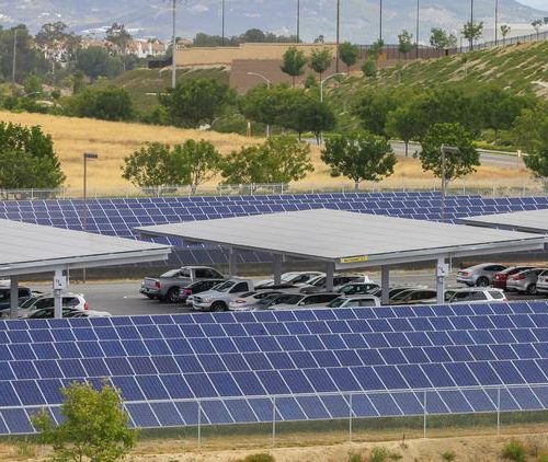 Cars parked under solar panel-covered carports in a large open parking lot, with grassy hills visible in the background.