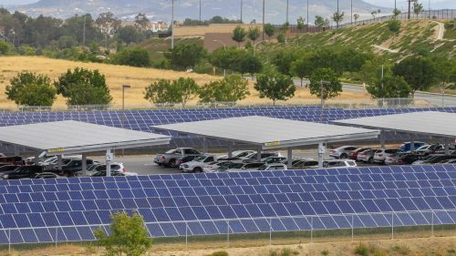Solar panels installed over a parking lot, providing shade to parked cars with a hilly landscape in the background.
