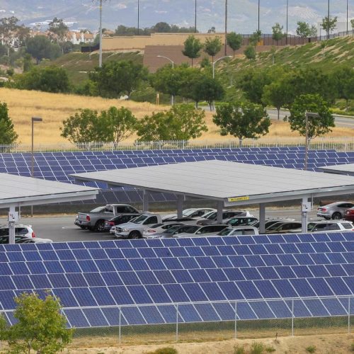 Solar panels installed over a parking lot, providing shade to parked cars with a hilly landscape in the background.