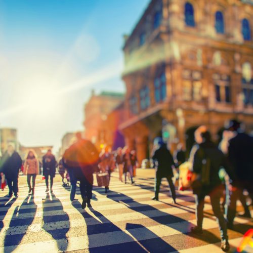 Blurred image of people crossing a busy urban street with sunlight flaring in the background, conveying a bustling city atmosphere.