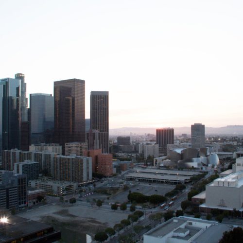 Aerial view of a downtown skyline at dusk, featuring tall skyscrapers and scattered buildings under a soft pink sky.
