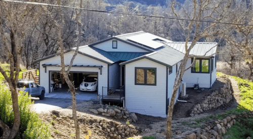 Modern house with a carport and a solar-paneled roof, surrounded by a stone-lined landscape and leafless trees.