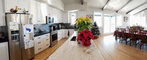 Panoramic view of a bright kitchen with white cabinets, a stainless-steel refrigerator, and a large dining area with a table set in the background.