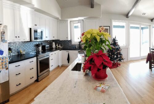 Panoramic view of a bright kitchen with white cabinets, a stainless-steel refrigerator, and a large dining area with a table set in the background.