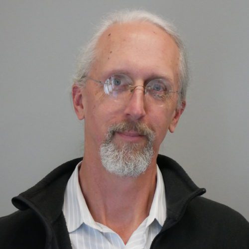 Headshot of a middle-aged man with grey hair, a beard and mustache, wearing glasses, a white shirt and a black jacket. he is looking directly at the camera.