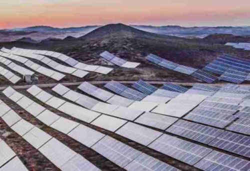 Expansive solar farm with rows of photovoltaic panels at dawn, set against a hilly landscape under a soft gradient sky.