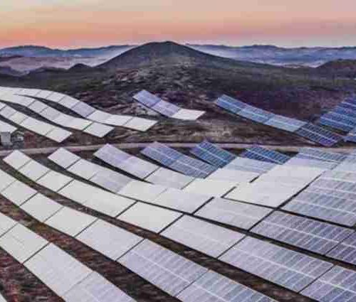 Expansive solar farm with rows of photovoltaic panels at dawn, set against a hilly landscape under a soft gradient sky.