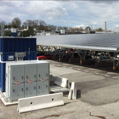 Solar panels installed over a parking lot with a shipping container and electrical equipment nearby under a cloudy sky.