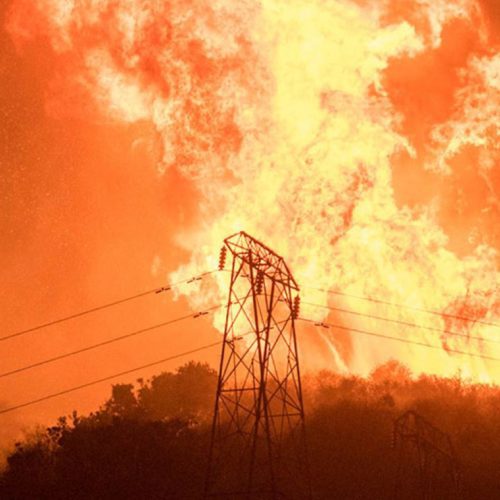 A massive wildfire engulfs a hillside, with towering flames rising behind power transmission towers connected to a community microgrid at dusk.
