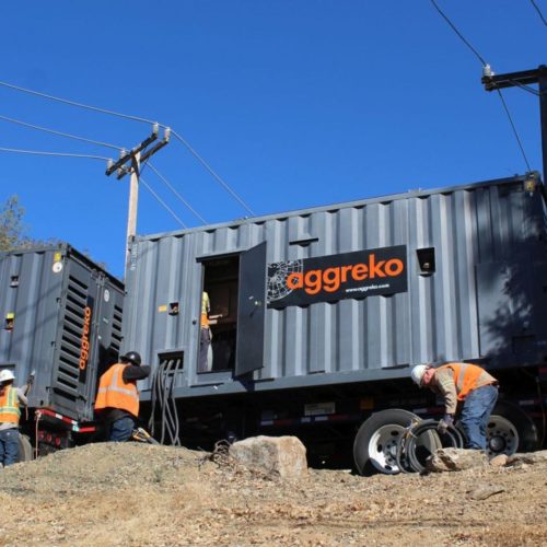Workers installing large aggreko generator units outdoors near electrical poles under a clear blue sky.