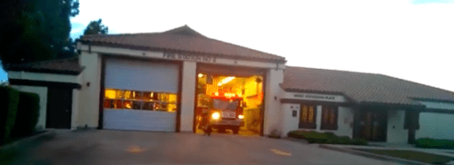 A fire truck exiting a fire station at dusk, with lights on and garage doors open.