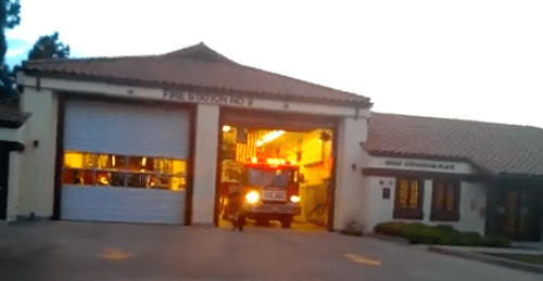 A fire truck exiting a fire station at dusk, with lights on and garage doors open.