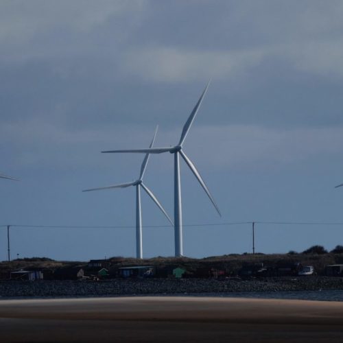 Three wind turbines on a coastal landscape under a cloudy sky, with a sandy beach in the foreground and a rocky shoreline.