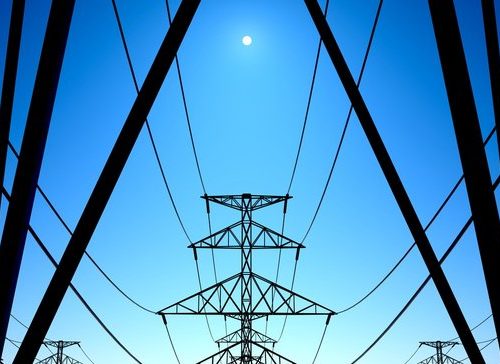 Silhouetted electricity pylons with crisscrossing wires against a clear blue sky, with a visible midday moon.