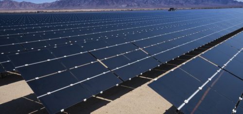 Expansive solar farm consisting of numerous rows of photovoltaic panels, set against a backdrop of distant mountains under a clear sky.