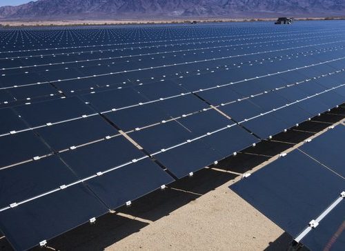 Expansive solar farm consisting of numerous rows of photovoltaic panels, set against a backdrop of distant mountains under a clear sky.