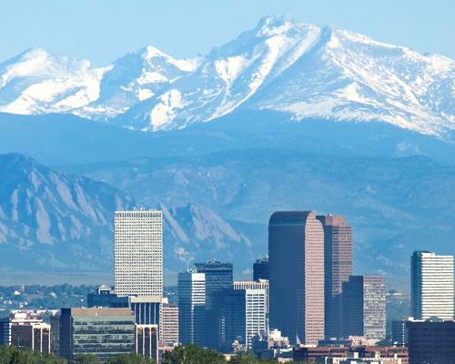 Panoramic view of a city skyline with modern buildings in the foreground and snow-capped mountains in the background under a clear blue sky.
