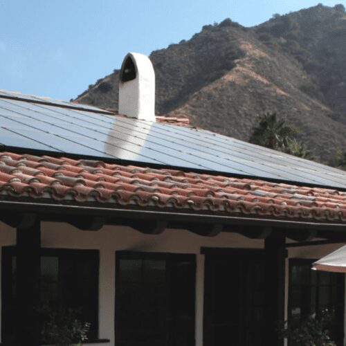 A house with a terracotta roof and solar panels, set against a backdrop of mountains, with white umbrellas visible in the foreground.