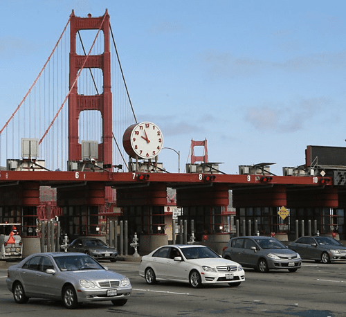 Traffic passing through a toll plaza with a large clock in front of the golden gate bridge.