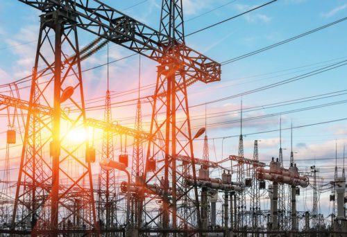 High-voltage power lines and transformers at a power station with the sun setting in the background, depicting energy infrastructure.