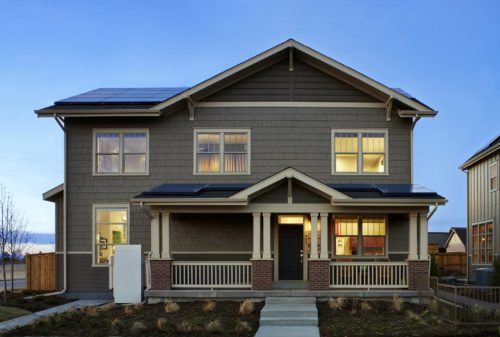 Modern two-story house with solar panels on the roof, visible at dusk with interior lights on and a clear sky.