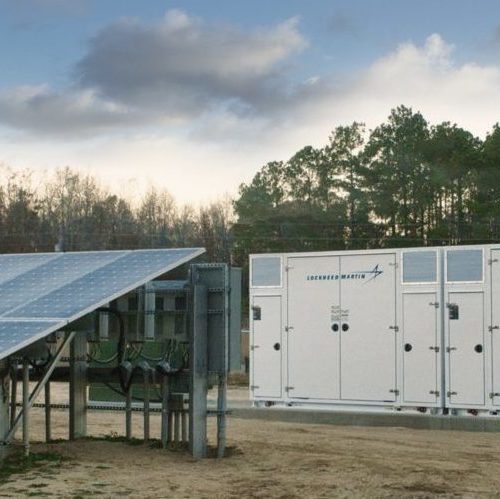 Solar panels adjacent to large electrical storage units in a field under a cloudy sky.