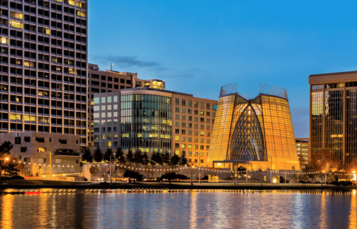 Twilight view of a modern cityscape featuring distinct glass buildings and reflections on a calm river.