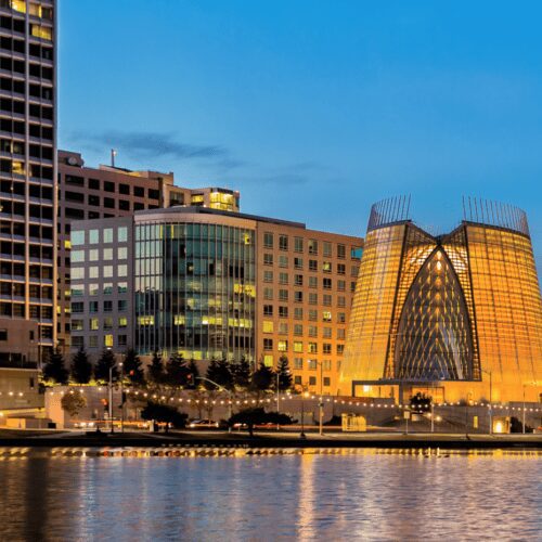 Twilight view of a modern cityscape featuring distinct glass buildings and reflections on a calm river.