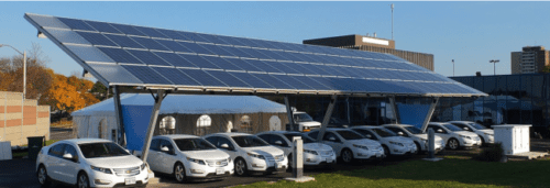 Solar panels installed over a carport sheltering parked cars on a sunny day, with buildings in the background.