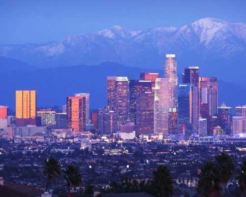 Downtown los angeles skyline at dusk with illuminated buildings, with snow-capped mountains in the background.