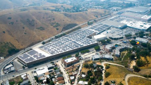 Aerial view of a large industrial complex with solar panels on the roofs, surrounded by roads and hills.