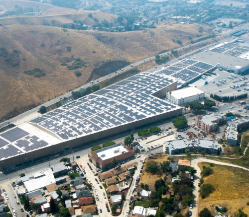 Aerial view of a large industrial complex with solar panels on the roofs, surrounded by roads and hills.