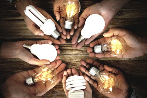 Several hands holding different types of light bulbs over a wooden table, representing diversity in lighting solutions.