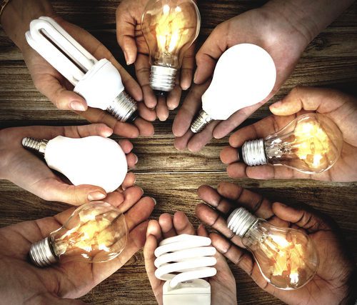 Several hands holding different types of light bulbs over a wooden table, representing diversity in lighting solutions.