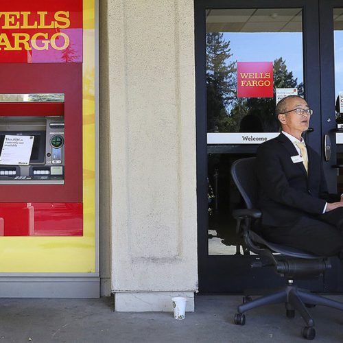 A security guard sitting beside a wells fargo atm entrance, wearing a suit and glasses, monitoring the surroundings.